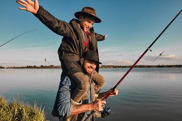 Obraz premium Son and father have fun while fishing. Father holds son on shoulders. Boy holds hands to sides. They smile. Background lake.