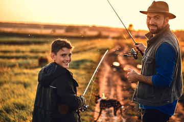 Father and son took fishing rods, fishing tackle and dog. They go on country road near lake for fishing. They smile and look at camera. Sunset. - Powered by Adobe