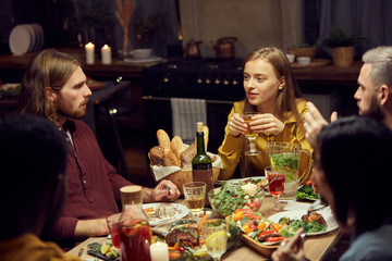 Group of modern young people enjoying dinner and wine together sitting at table in dimly lit room, copy space