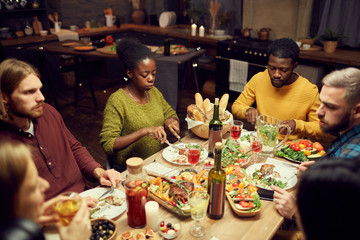 Multi-ethnic group of friends enjoying dinner and wine together sitting at table in dimly lit room, copy space