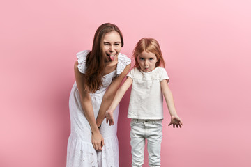 crazy mother sticking her tongue while her child is being puzzled, kid shrugging her shoulder. close up photo, entertainment. isolated pink background