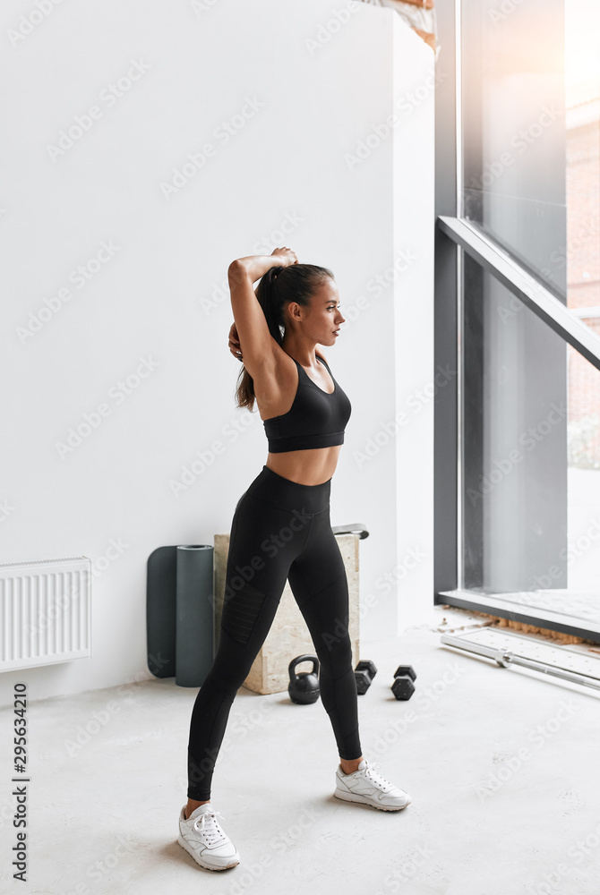 Wall mural athletic female doing exercises standing at gym. dressed in black clothes. background window