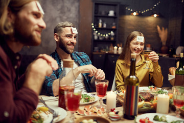 Group of cheerful young people playing guessing game while sitting at table during dinner party in dark room, copy space