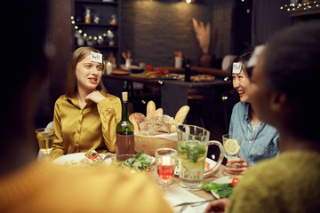 Portrait of two cheerful young women playing guessing game while enjoying dinner party with friends sitting at table in dark room, copy space