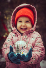 Child hold transparent glass ball with christmas tree and house inside.
