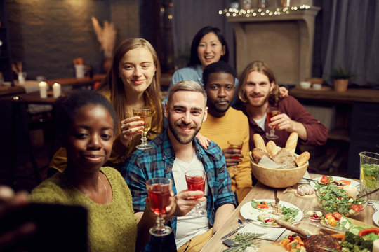 Multi-ethnic group of friends taking selfie photo via smartphone while enjoying dinner party at home, copy space