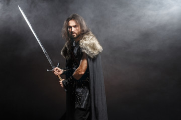 Man dressed in medieval armor and raincoat with longs word over smoke background. Courage fantasy...