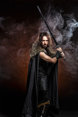 Fototapeta premium Man dressed in medieval armor and raincoat with longs word fighting against enemy. Courage fantasy warrior knight with long hair concept historical photo