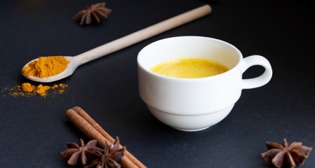Golden (turmeric) milk in a white cup on the dark background. A spoon with turmeric powder.  Cinnamon and anise.