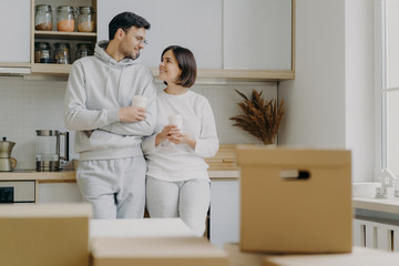 Image of cheerful woman and man have pleasant talk during coffee break, wear casual clothes, move in new apartment, pose with modern furnished kitchen, unpack cardboard boxes with household stuff