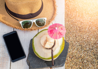 Fresh coconut drink and summer accessories, sunglasses, hat, mobile phone on white wood table with sunlight
