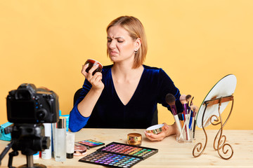 Front view of pretty young blogger girl frowning in disgust, sitting at dressing table full of diverse makeup products with dissapointed face expression, probably face cream is expired.