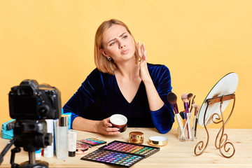 Thoughtful female makeup artist applying new face cream, has focused and contemplative expression, recording new podcast with tips and beauty hacks for followers. Massmedia and freelance concept.