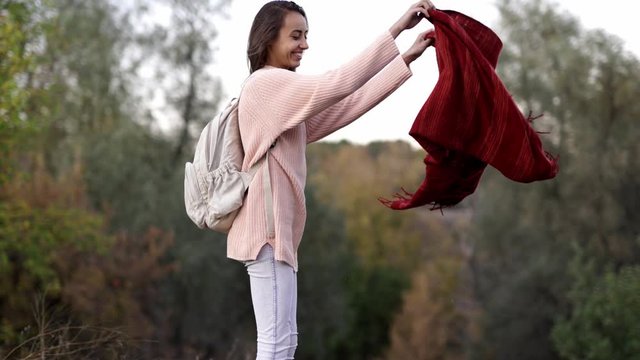 pretty smiling Woman in pink knitted sweater with small backpack shakes the blanket after picnik in the autamn forest and going out. girl hipster walking outdoors on colorful fall park background, 4k