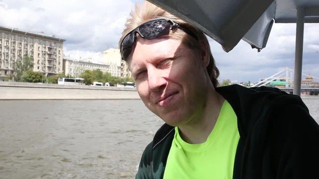 close-up of young man in sunglasses wearing green sweatshirt, blinking his eyes against water and resting on handrail, floating on Moscow river in boat on sunny day. water transport tourist trip