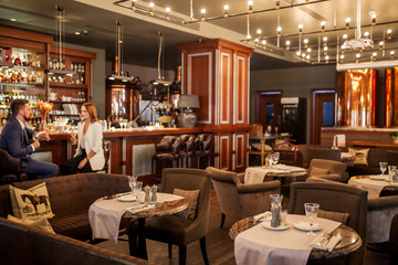 Large beautiful restaurant with round tables, and soft chairs, room lighted brightly by many small lamps, young couple sits and drinks fragrant white wine at bar counter