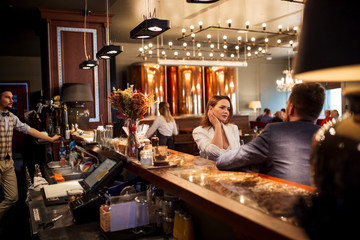 Naklejka premium Attractive good shaped woman dressed in white clothes, touching neck thoughtfully sitting at bar counter together with handsome friend at restaurant, side view