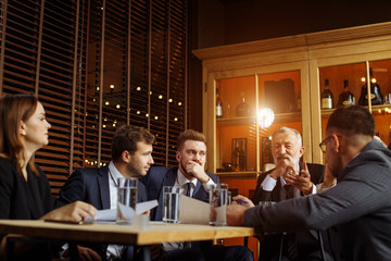 Young businessmen siting at wooden table and listening attentively to old beard man, nice looking leader enthusiastically speaking and gesturing in the office of company