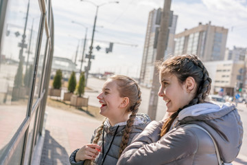 two girlfriends teenagers in warm gray jackets laugh looking at glass reflection of shop window...