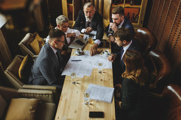 Business people in formal suits sitting at light-coloured conference table, discussing documents. Bearded man holding out hand and pointing to an important clause of business contract