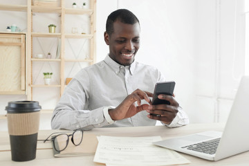 Close-up of african businessman sitting at table in office and smiling while using smartphone