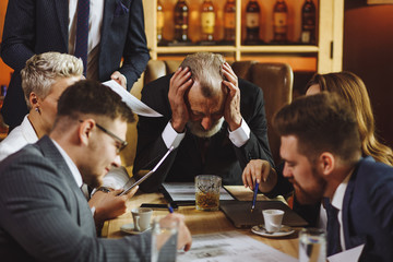 Business leader closed ears with hands and carefully reads important contract trying to concentrate, young partners sit at wooden table vigorously discussing upcoming changes