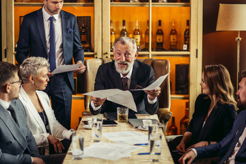 Rather old gray-haired man with beard enthusiastically telling something to business partners, holding documents in his hands, looking directly at camera with open mouth