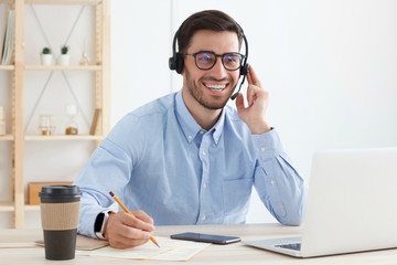 Smiling customer support service operator with hands-free headset working in office, using laptop