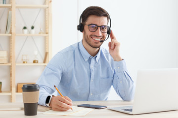 Portrait of smiling male customer service worker, sitting with laptop at desk in white modern office