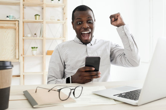 Young african american shocked business man using phone and making winner gesture with fist