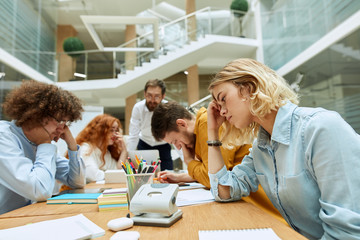 Dreamy young woman in light blue shirt, hold head, touches forehead with fingers, looks aside, thinks over work related issues in modern coworking office, shot from below