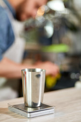 Metal coffee pot stands on kitchen scales, blurred background of working barman behind bar counter, close up, professional concept