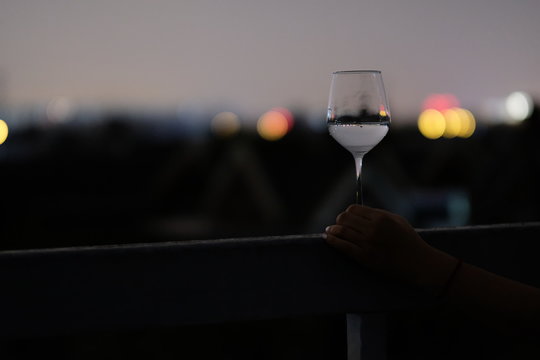 One Hand Holding A Glass Of White Wine On Handrail. Dark Night Sky And Blur Colorful City Lights Background