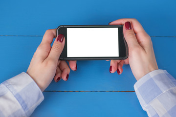 Mockup image: woman looking at black smartphone with white blank screen on blue table. Close up view of woman hands with smart phone mobile device. Mock up, copyspace, template and technology concept