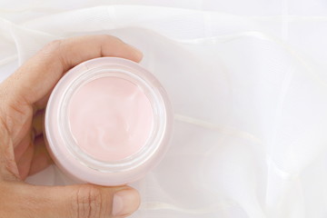 Fototapeta na wymiar Top view of close up woman hand holding pink cream jar on white fabric background, have copy space