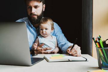 Father with little baby on knees making notes in notebook, looking at laptop. Young man holding...