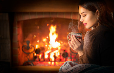 Beauty young Christmas woman sitting near fireplace in dark room at home and drinking hot beverage from a big cup. Beauty girl Enjoying tea, coffee or hot chocolate in a cozy room. Xmas scene - Powered by Adobe