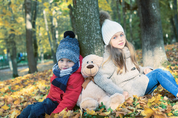 Little boy and girl with Teddy Bear friend sitting on the ground in the autumn park on sunset, nice back light.