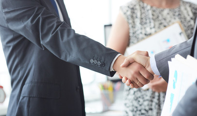 Business people in suits shaking hands, finishing up a meeting. Male hands in handshake closeup with female colleague in background.