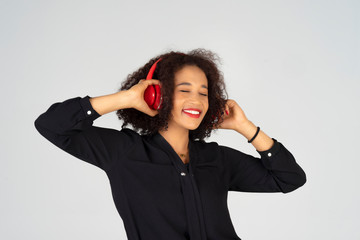 Young African woman listening music from wireless or Bluetooth red headphones, control online track from smartphone while standing in a grey background. Feel relaxation, happiness and cheerful.