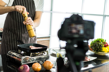 Food blogger making pork steak for online teaching, sprinkle pepper on top in home kitchen with ingredients on table. Taking video by using camera and explain step to make a delicious meal.