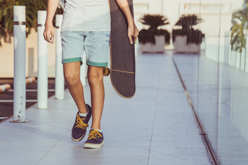 Trendy child walking in a modern city context. Young boy with skateboard in hand walks alone. Close up view of young male legs strolling in a modern mall. Teen at the shopping center. Youth concept.