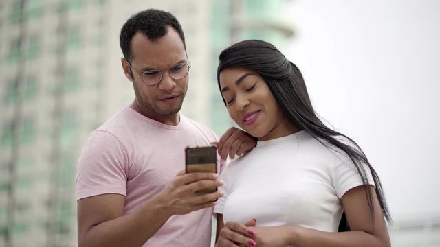 Smiling multiracial couple talking and looking at smartphone. Front view of cheerful young people with smartphone. Communication and technology concept