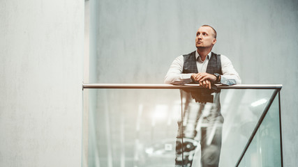 An adult caucasian pensive man entrepreneur is leaning on the chromium fence of a glass balcony in...