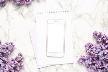 Mobile phone, notebook and purple lilac flowers on marble background, simple business composition with spring flower