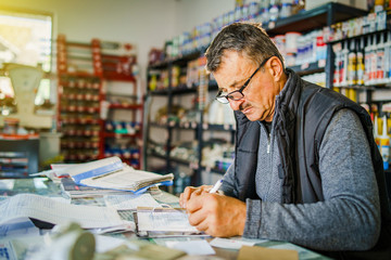 Senior man entrepreneur at his office store by the table accounting check bill calculation receipt making invoice for selling goods salesman at warehouse depot storehouse