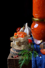 Adjika on a slice of rye bread and in a glass jar surrounded by the ingredients on the edge of the wooden table