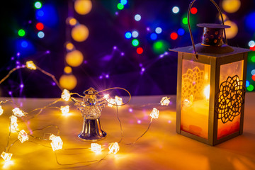 Candlestick with glowing candle, glowing lights and gold angel in tne centre are on the shiny table/background.There are different colors lights on the background. Merry Christmas. Happy New Year 2020