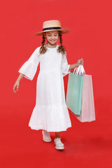 Cute little girl smiling, wear in dress and hat, holdings in hands shopping bags, on red background. Vertical view.