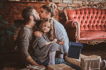 Surrounded by christmas gifts, mother and father are kissing, while holding their little daughter.